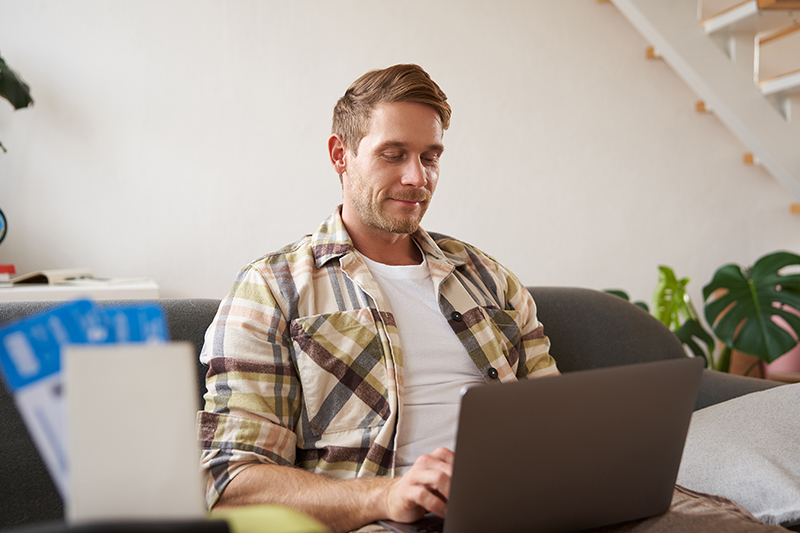 Portrait of handsome young man, has two tickets and passport, goes online on laptop, booking hotel and planning his holiday, sitting in living room.